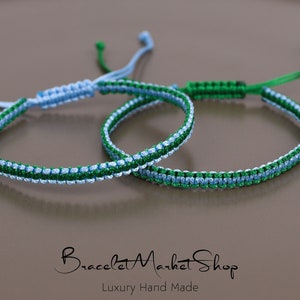 May include: Two friendship bracelets, one blue and green, the other green and blue, are knotted together. The bracelets are made with a woven pattern and have a simple knot closure.