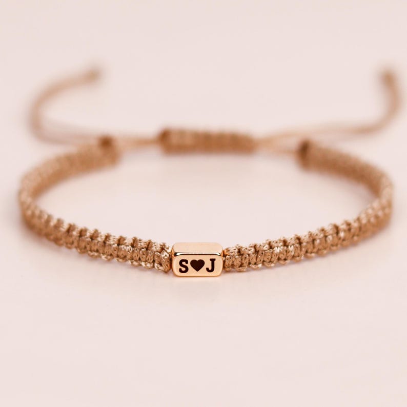 May include: A gold-colored macrame bracelet with a sliding knot closure. The focal point is a small, rectangular gold bead with the letters "S" and "J" separated by a heart symbol. The bracelet is adjustable.