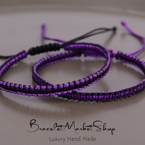 May include: Two friendship bracelets, one black and purple, the other purple and black, are knotted together on a light brown surface. The bracelets are made with a woven cord.