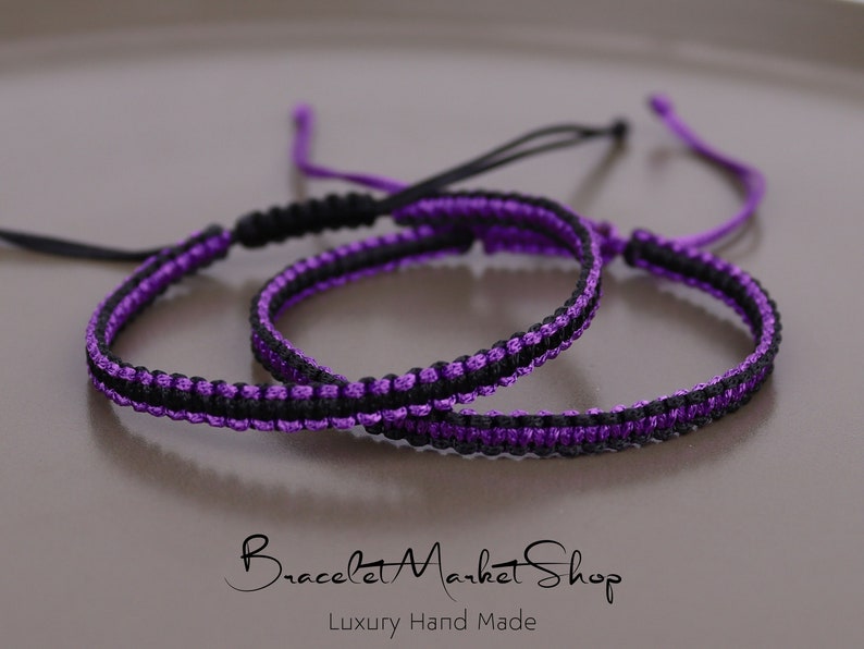 May include: Two friendship bracelets, one black and purple, the other purple and black, are knotted together on a light brown surface. The bracelets are made with a woven cord.