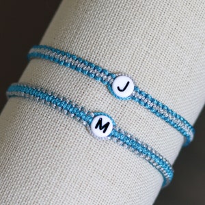 May include: Two friendship bracelets with blue and silver threads. The bracelets have white beads with the letters "J" and "M" on them.