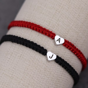 Pode incluir: Duas pulseiras de amizade com pingentes em forma de cora&ccedil;&atilde;o. A pulseira de cima &eacute; vermelha e tem a letra "A" no pingente. A pulseira de baixo &eacute; preta e tem a letra "J" no pingente.