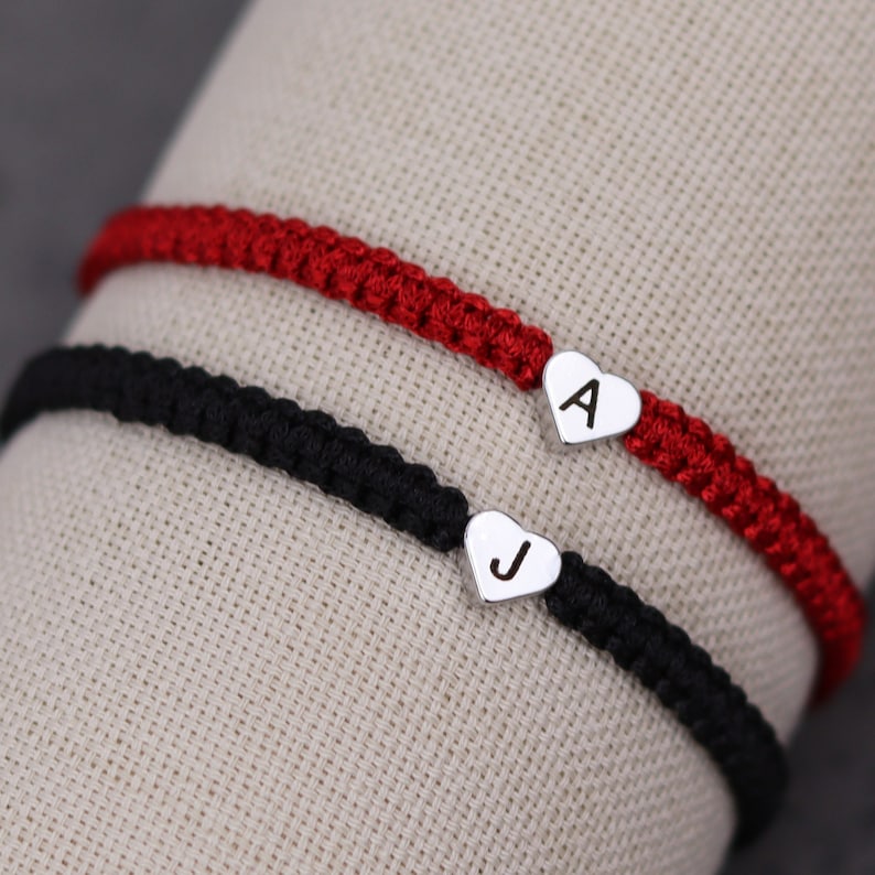 Pode incluir: Duas pulseiras de amizade com pingentes em forma de cora&ccedil;&atilde;o. A pulseira de cima &eacute; vermelha e tem a letra "A" no pingente. A pulseira de baixo &eacute; preta e tem a letra "J" no pingente.