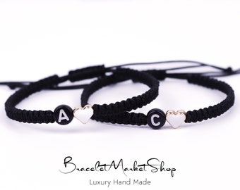 May include: Two black friendship bracelets with white heart charms and black letter beads. The bracelets spell out the letters "A" and "C".