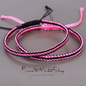 May include: Two friendship bracelets, one pink and one black, are woven together. The bracelets are made with a knotting technique and have a simple, elegant design.