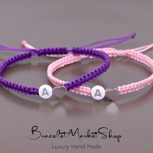 Op de afbeelding: Twee vriendschapsarmbanden met een hartbedel en een letter 'A' bedel. De armbanden zijn paars en roze.