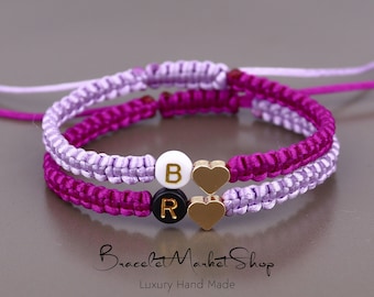 May include: Two matching friendship bracelets with purple and pink braided cords. The bracelets have gold heart charms and letter beads that spell "BR".