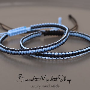 May include: Two friendship bracelets, one black and light blue, the other light blue and black, are knotted together. The bracelets are made with a woven cord and have a simple design.