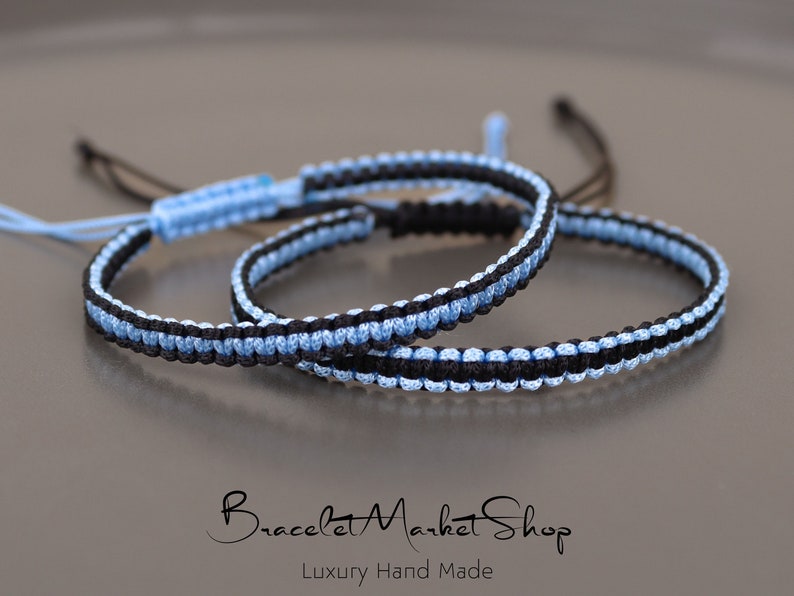 May include: Two friendship bracelets, one black and light blue, the other light blue and black, are knotted together. The bracelets are made with a woven cord and have a simple design.