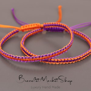 May include: Two friendship bracelets, one orange and purple, the other purple and orange, are knotted together. The bracelets are made with a simple, woven pattern.