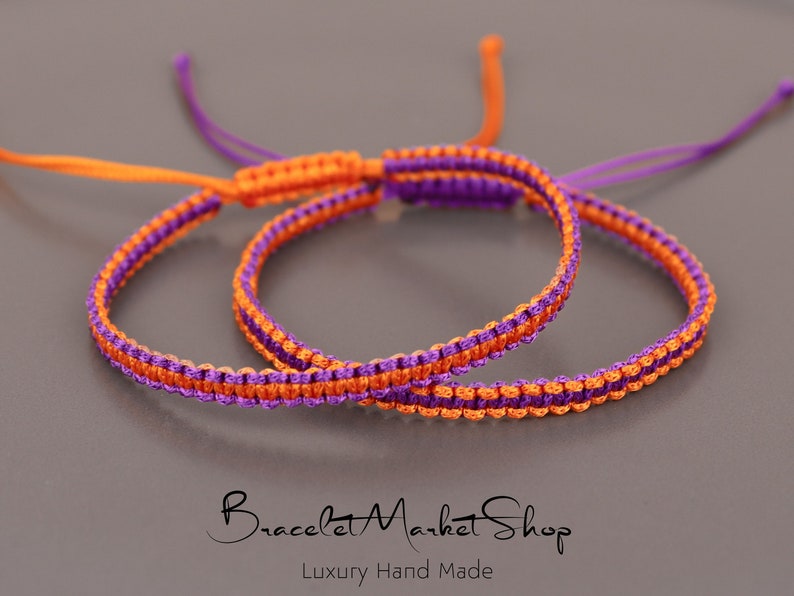 May include: Two friendship bracelets, one orange and purple, the other purple and orange, are knotted together. The bracelets are made with a simple, woven pattern.