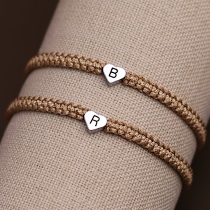 Pode incluir: Duas pulseiras de amizade marrons com pingentes em forma de cora&ccedil;&atilde;o. Os pingentes s&atilde;o prateados e t&ecirc;m as letras "B" e "R" gravadas.