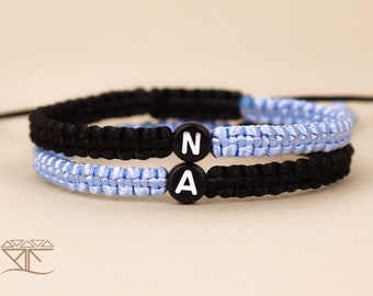 Può includere: Due braccialetti dell'amicizia con cordini neri e blu chiaro. I braccialetti hanno perline nere con lettere bianche che compongono la parola "NA".