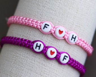 May include: Two friendship bracelets with pink and purple cords. The pink bracelet has white beads with the letters "F" and "H" and a red heart bead. The purple bracelet has white beads with the letters "H" and "F" and a red heart bead.