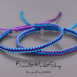 May include: Two friendship bracelets, one blue and purple, the other purple and blue, are knotted together. The bracelets are made with a simple, woven pattern.