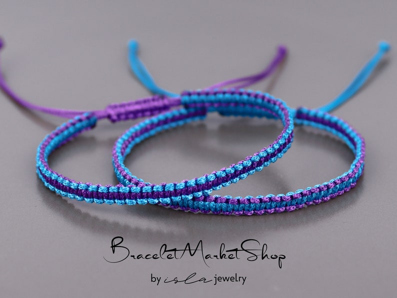 May include: Two friendship bracelets, one blue and purple, the other purple and blue, are knotted together. The bracelets are made with a simple, woven pattern.