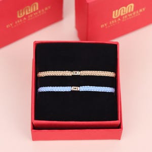 May include: Two personalized bracelets with initials in a red gift box. One bracelet is tan, the other is light blue. The bracelets are displayed on a black velvet insert. The box has the text "BY ISLA JEWELRY".