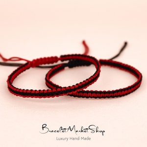 May include: Two red and black friendship bracelets with a knot at the end. The bracelets are made of thin cord and are layered on top of each other.