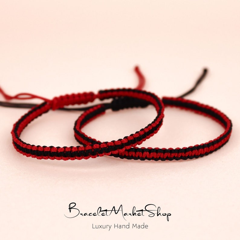 May include: Two red and black friendship bracelets with a knot at the end. The bracelets are made of thin cord and are layered on top of each other.
