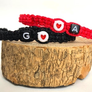 May include: Two friendship bracelets, one black and one red, with a heart charm and letter beads. The black bracelet has a "G" bead and the red bracelet has an "A" bead.