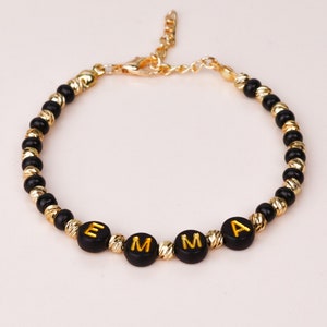 May include: A black beaded bracelet with a gold clasp and gold lettering spelling out the name "EMMA".