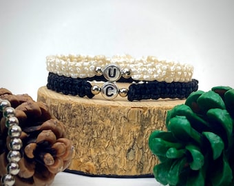 May include: Two friendship bracelets with the letters "U" and "C" on silver beads. The bracelets are made with black and white cord and are stacked on a wooden surface.