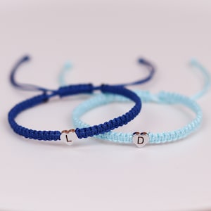 Pode incluir: Duas pulseiras de amizade com pingentes em forma de cora&ccedil;&atilde;o. Uma pulseira &eacute; azul e a outra &eacute; azul claro. A pulseira azul tem a letra "L" no pingente e a pulseira azul claro tem a letra "D" no pingente.