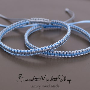 May include: Two braided friendship bracelets in blue and silver. The bracelets are tied together and are lying on a gray surface.