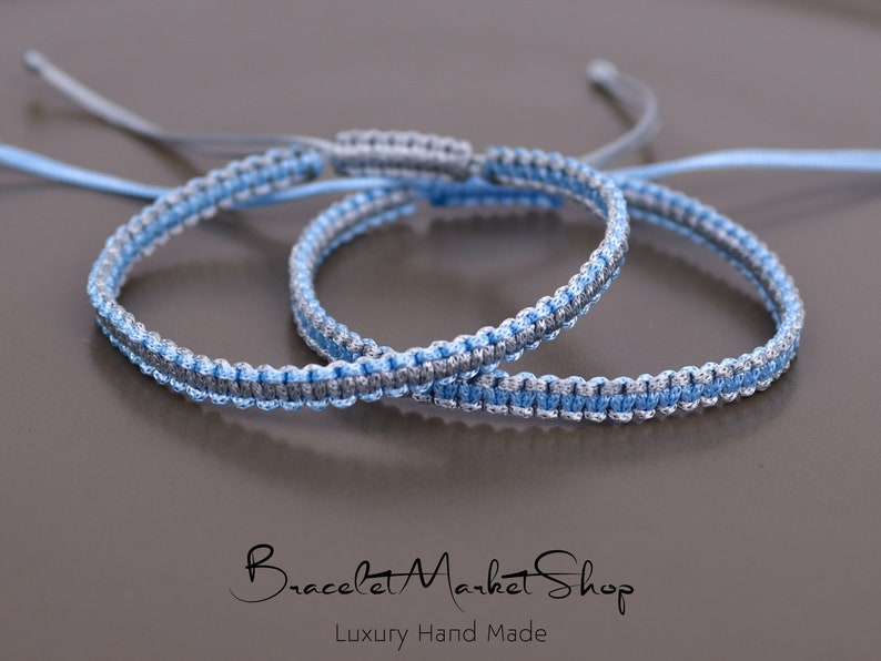 May include: Two braided friendship bracelets in blue and silver. The bracelets are tied together and are lying on a gray surface.