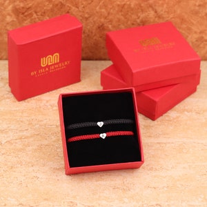 Pode incluir: Duas pulseiras de cord&atilde;o vermelho com pingentes de cora&ccedil;&atilde;o prateados e as letras J e A. As pulseiras est&atilde;o em uma caixa vermelha com interior preto. A caixa tem o logotipo "U&M BY ISLA JEWELRY" na parte superior.