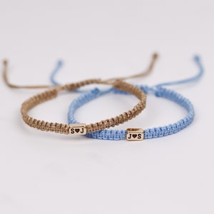 May include: Two macrame bracelets. One is tan with a gold-colored rectangular bead with the letters "S" and "J" and a heart symbol. The other is light blue with a similar bead with "J" and "S" and a heart symbol.