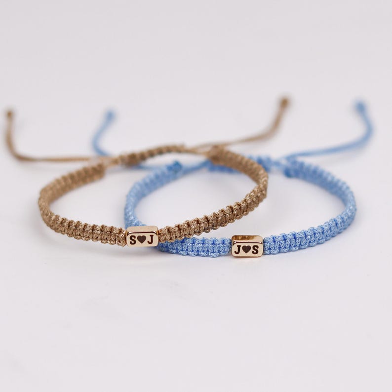 May include: Two macrame bracelets. One is tan with a gold-colored rectangular bead with the letters "S" and "J" and a heart symbol. The other is light blue with a similar bead with "J" and "S" and a heart symbol.