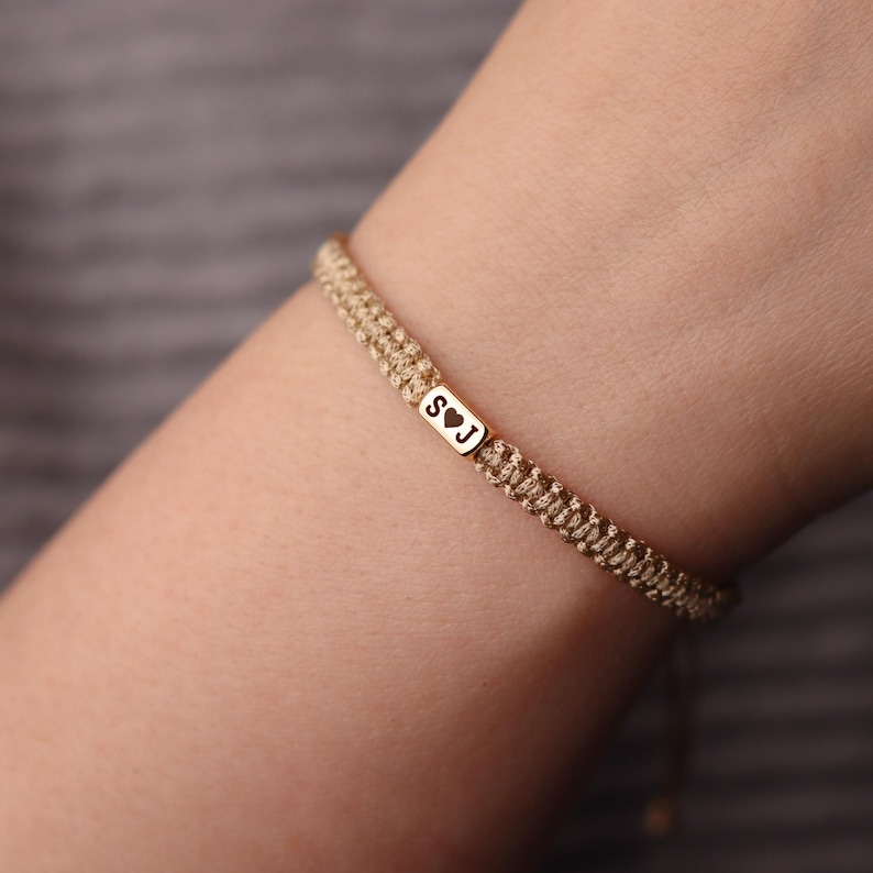 May include: A woven beige bracelet with a small gold-colored rectangular charm. The charm has the letters "S" and "J" with a heart symbol between them. The bracelet is on a person's wrist.