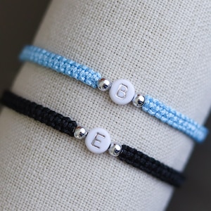 Puede incluir: Dos pulseras de macramé, una azul claro y otra negra, cada una con una cuenta circular blanca con una letra. La pulsera azul claro tiene la letra "B", y la pulsera negra tiene la letra "E". Ambas pulseras tienen cuentas plateadas.
