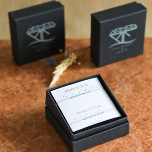 May include: Two black boxes with a silver diamond logo and the text "Make It Fun" in braille on a white card inside. The braille is represented by small turquoise beads.