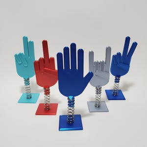 May include: Five colorful hand figurines on spring-loaded bases. The hands are in different poses, including peace signs, a middle finger, and a rock and roll sign. The bases are blue, red, silver, and blue.
