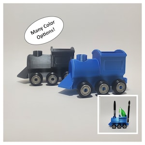 May include: Two 3D printed toy train engines, one blue and one black, with wheels. The blue engine has a compartment for holding pens or pencils. The text "Many Color Options!" is in a white circle above the trains.
