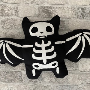 May include: A black bat-shaped cushion featuring a white skeleton design. The cushion has outstretched wings and a detailed skeleton print on the front. This Halloween-themed decorative cushion is made of soft fabric.