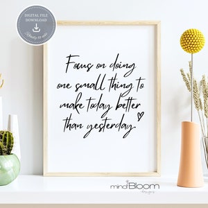 Puede incluir: Una impresión de arte de pared imprimible con fondo blanco y texto negro que dice "Focus on doing one small thing to make today better than yesterday." El texto está en una fuente de escritura a mano y hay un pequeño símbolo de corazón al final del texto. La impresión está enmarcada en un marco de madera claro y está sentada en un estante blanco con una planta de cactus, un jarrón de flores secas y un jarrón de flores amarillas.