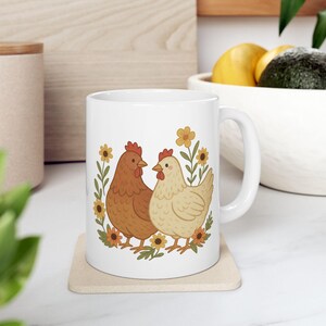 May include: White ceramic mug featuring a charming illustration of two chickens, one brown and one cream-colored, surrounded by yellow flowers and green leaves. The mug has a curved handle and is set on a beige coaster.