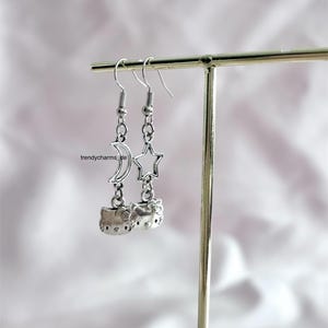 Könnte beinhalten: Silberfarbene Hängeohrringe mit Hello Kitty-Design. Ein Ohrring hat einen Mond-Anhänger, der andere einen Stern. Die Ohrringe sind an einem Metallhaken und einer Kette befestigt.