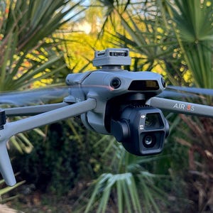 May include: A grey drone with a camera and the text "AIR 2S" on the side. The drone is in flight against a background of green foliage.