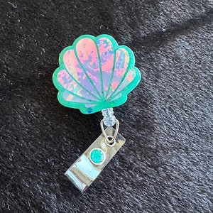 May include: A green iridescent seashell-shaped badge holder with a clear plastic retractable cord and a silver clip with a green gem.