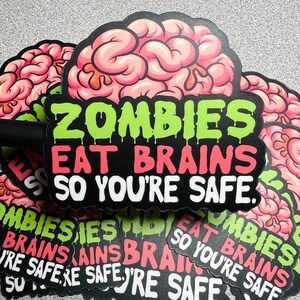 Op de afbeelding: Een stapel stickers met een roze en witte hersenen afbeelding en de tekst "Zombies Eat Brains So You're Safe."