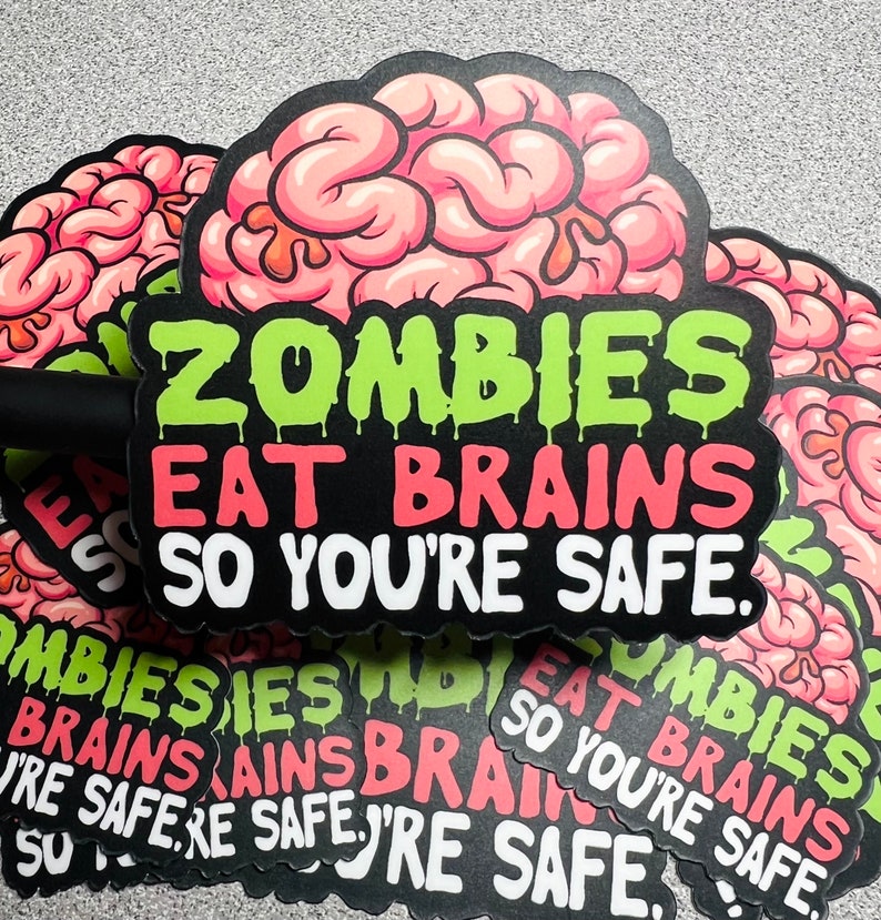 Op de afbeelding: Een stapel stickers met een roze en witte hersenen afbeelding en de tekst "Zombies Eat Brains So You're Safe."