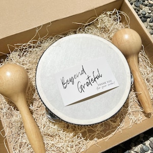 Könnte beinhalten: Ein weißes Tamburin mit schwarzem Rand, zwei Holzmaracas und eine Karte mit der Aufschrift "Beyond Grateful" mit dem Text "Thank you for your order!" in einem braunen Karton, gefüllt mit zerkleinertem Papier.