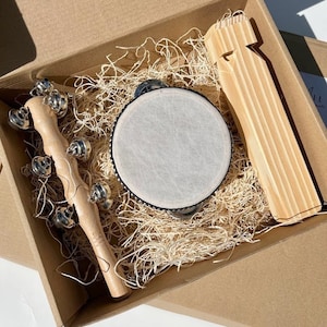 Könnte beinhalten: Eine braune Pappbox gefüllt mit Holzmusikinstrumenten. Die Box enthält ein Tamburin, eine Reihe von Glöckchen an einem Holzstab und einen Holzklatscher.