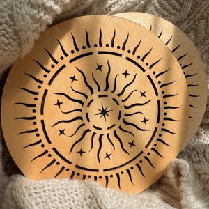K&ouml;nnte beinhalten: Zwei Holzuntersetzer mit einem Sonnen- und Sternenmuster. Die Untersetzer sind hellbraun mit einem schwarzen, ge&auml;tzten Sonnenmuster. Das Sonnendesign hat gebogene Strahlen, Sterne und einen zentralen Stern. Die Untersetzer sind kreisf&ouml;rmig.