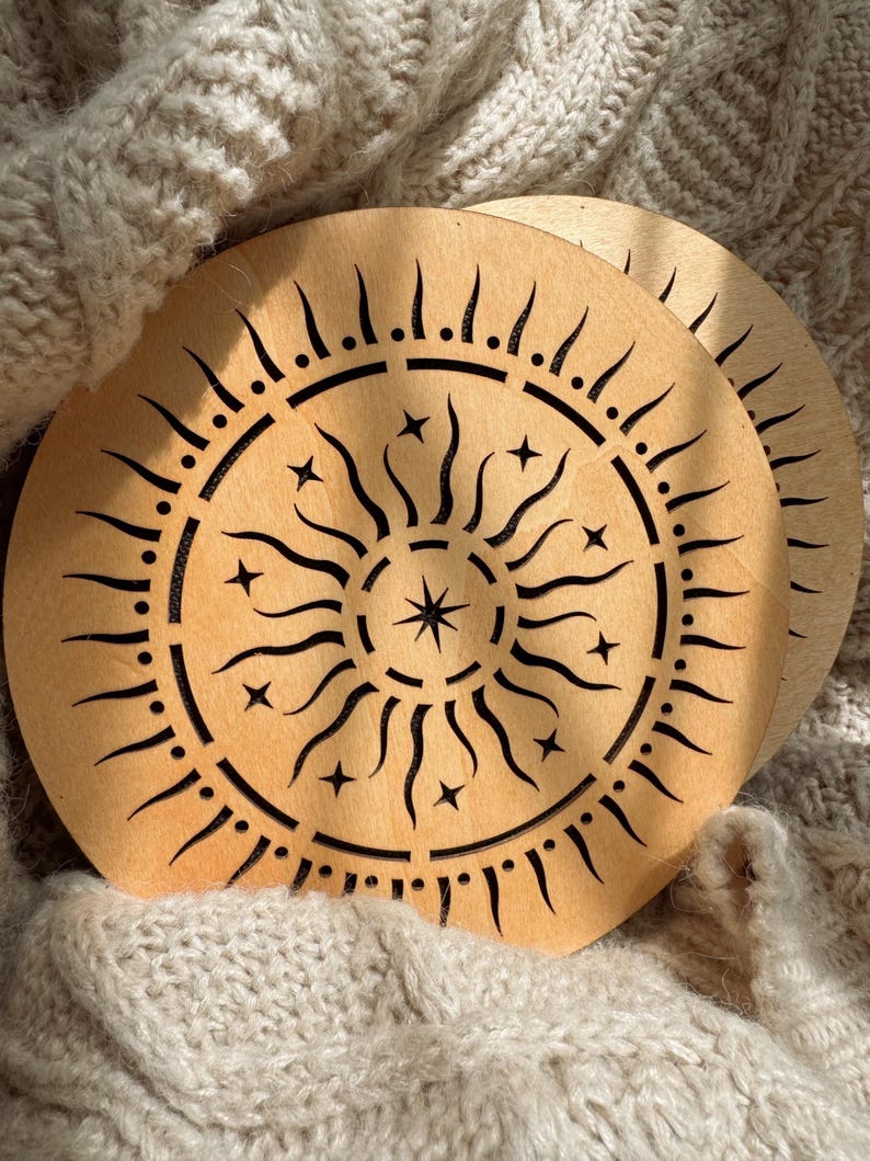 K&ouml;nnte beinhalten: Zwei Holzuntersetzer mit einem Sonnen- und Sternenmuster. Die Untersetzer sind hellbraun mit einem schwarzen, ge&auml;tzten Sonnenmuster. Das Sonnendesign hat gebogene Strahlen, Sterne und einen zentralen Stern. Die Untersetzer sind kreisf&ouml;rmig.