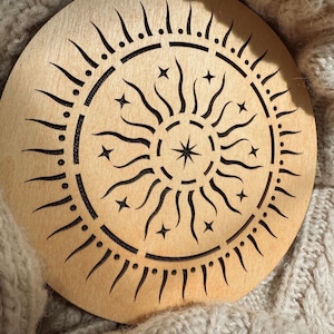 K&ouml;nnte beinhalten: Ein runder Holzuntersetzer mit einem Sonnen- und Sternenmuster. Die Sonnenstrahlen und Sterne sind ausgeschnitten, wodurch ein detailliertes Muster entsteht. Der Untersetzer ist hellbraun und liegt auf einer beigefarbenen Strickoberfl&auml;che.